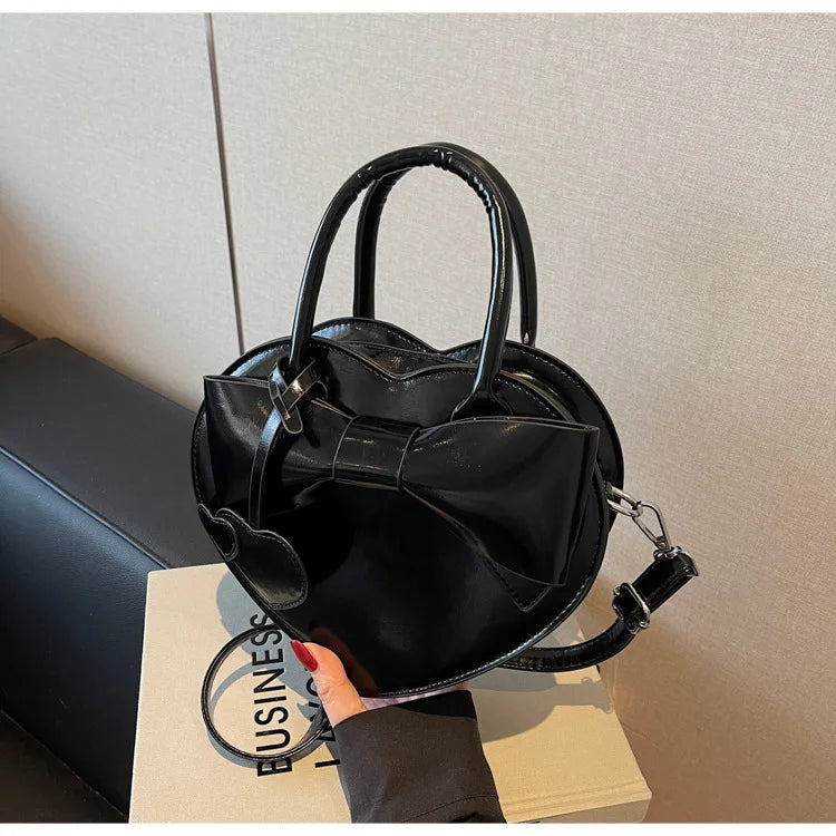 Black carauana-lovely-shoulder-bag-heart with a bow detail held by a hand on a neutral background